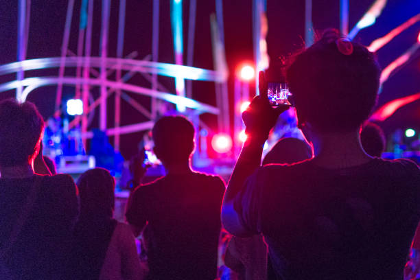 Ko Lanta, Krabi, Thailand - March 8, 2018: One unrecognisable young person of Asian ethnicity is recording a music concert on their mobile telephone.  They are holding the phone in front of their face and watching the activities through it's screen.  Several other people are standing nearby.  In the background a generic unrecognisable musician, performing on a stage, with brightly coloured lights flashing.  This is a common site these days, where people watch an event through their mobile telephone, instead of enjoying the moment.  Location is a free public concert held at Ko Lanta, Krabi, Thailand.  The event is on the public street, where the public have a right of access and no money is charged to watch or participate.